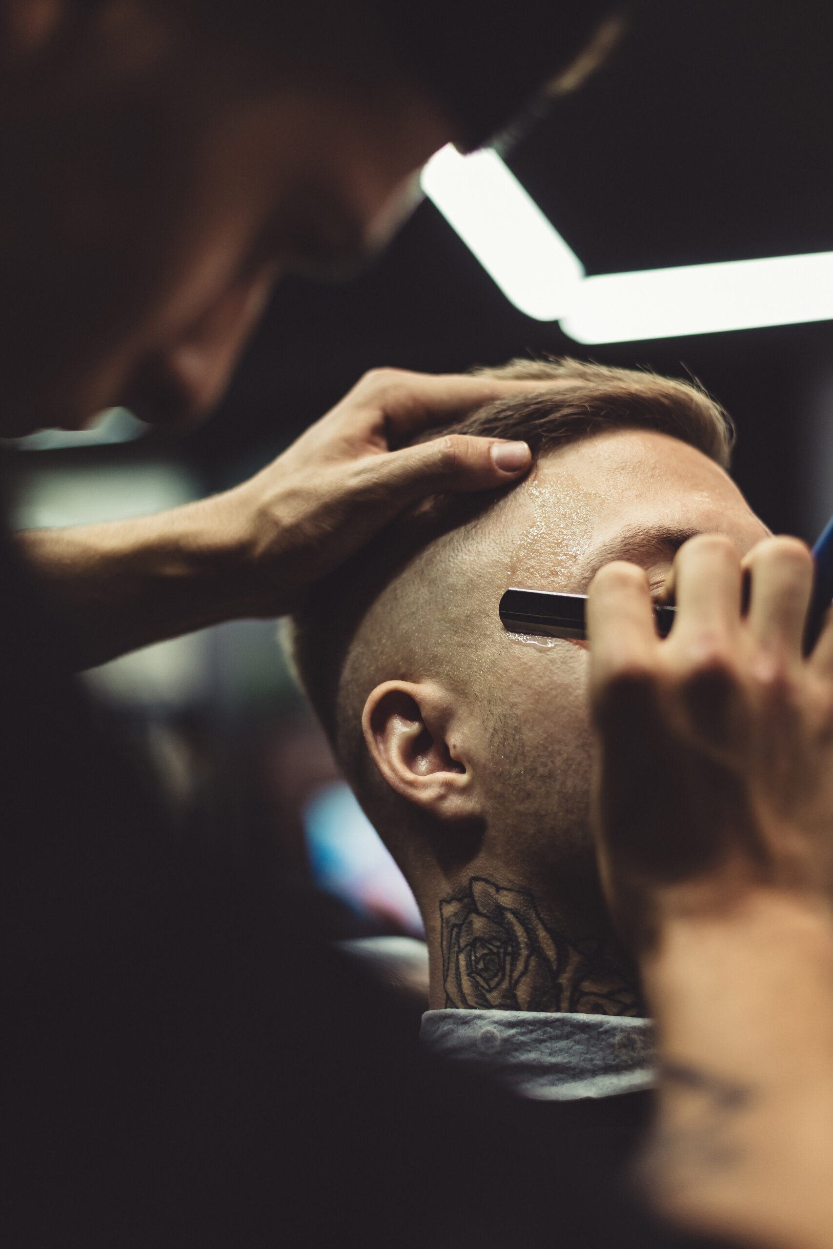Anonymous barber shaving stylish tattooed man with razor in barbershop.